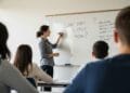 Teacher writing big, open-ended questions on a classroom whiteboard while students jot down notes