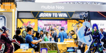 A cosplayer with blue and green braided hair in a futuristic outfit holding a yellow smartphone, posing at a tech event.