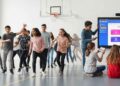 A vibrant, dynamic image of a diverse group of students actively engaged in a modern physical education setting. Some are participating in a sport (e.g., basketball or soccer) on one side, while others are interacting with tablets or smartboards on the other, displaying engaging quiz content. The setting is a brightly lit, contemporary gym or open space, emphasizing the integration of physical activity with digital learning tools. The overall mood is energetic, inclusive, and forward-thinking, capturing the essence of Gimkit transforming PE and health education.