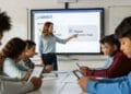 Photo-realistic image of a middle school classroom where a smiling teacher is setting up a Gimkit game on an interactive smartboard, while students actively participate on tablets and smartphones, demonstrating high engagement with the educational game platform.